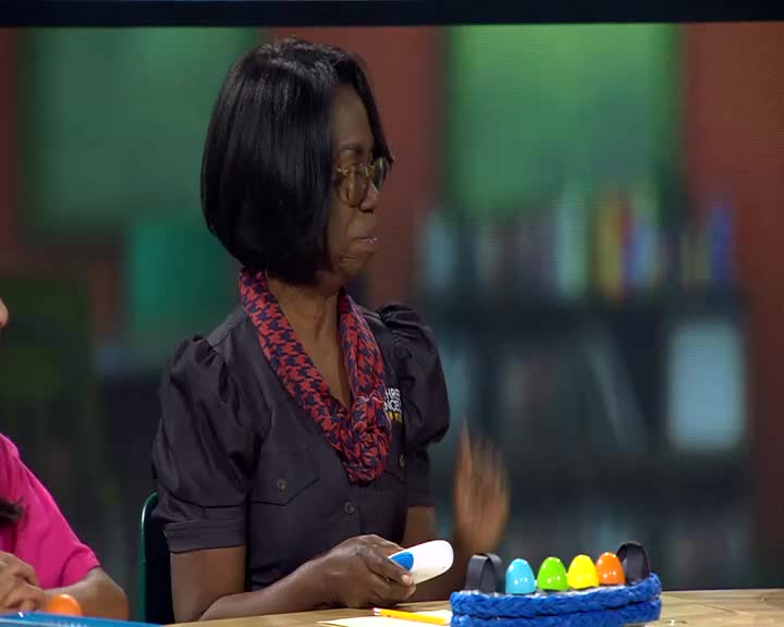 A woman with glasses gestures with her right hand over a colorful toy xylophone. She holds a white and blue marker in her left hand.