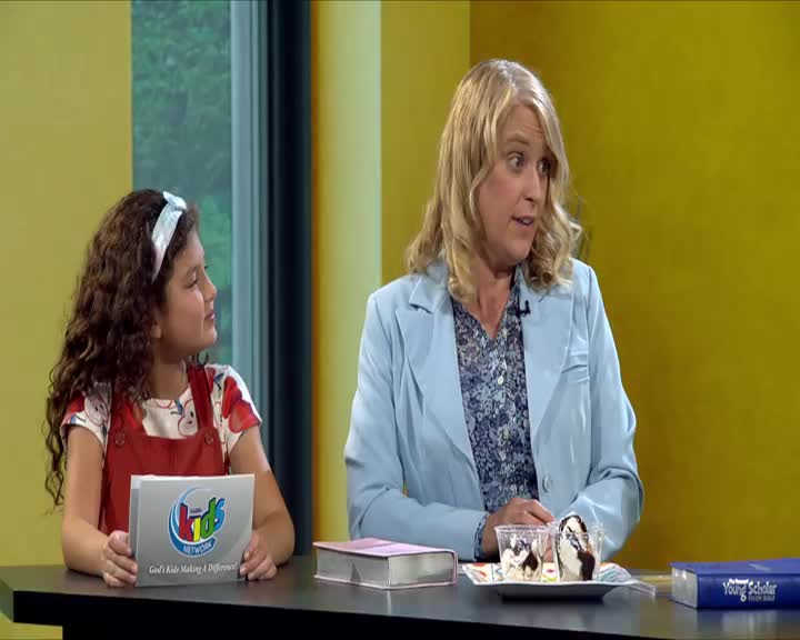 A young girl holds up a card with the 3ABN Kids Network logo. Beside her, a woman in a light blue blazer speaks animatedly.