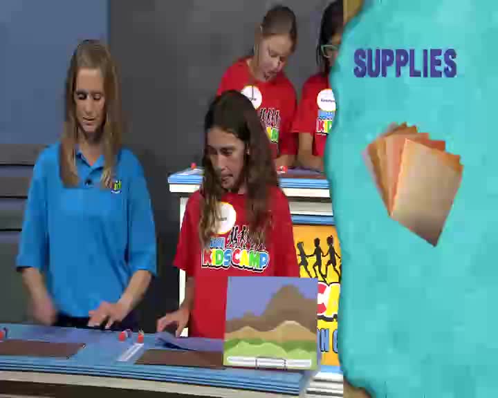 A woman in a blue shirt guides two girls in red shirts as they work on a craft project. They're gathered around a table with art supplies, likely for a 3ABN Kids program.