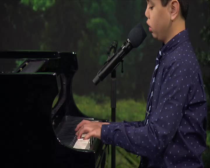 A young boy in a blue patterned shirt and tie plays the piano, his fingers moving across the keys. He sings into a microphone positioned in front of him, a backdrop of green trees behind him.