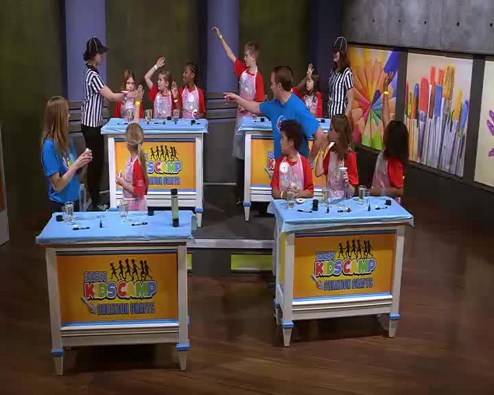Kids stand at their stations, some with hands raised, as a woman in a referee shirt directs them. A man points towards one of the children, and the 3ABN Kids Camp banner is visible on the front of their tables.