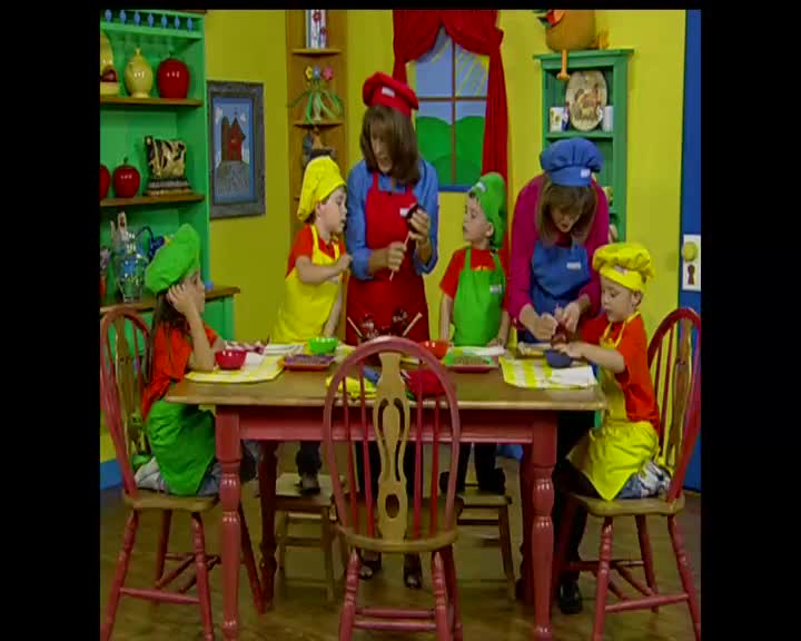 A group of children in colorful chef hats and aprons are gathered around a table, intently focused on their tasks. A woman in a red apron guides them, her hands busy with something small.