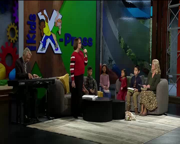 A woman in a red and white striped shirt sings into a microphone, while a man plays a keyboard to her left. Children sit on a couch to her right, listening intently.