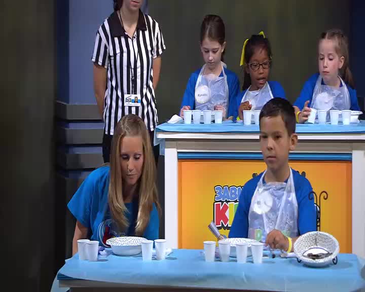 A young boy stares ahead, his brow furrowed in concentration, while a woman with blonde hair leans forward, her gaze fixed on the table in front of her. Behind them, three girls stand at a counter, each with a row of small cups before them.