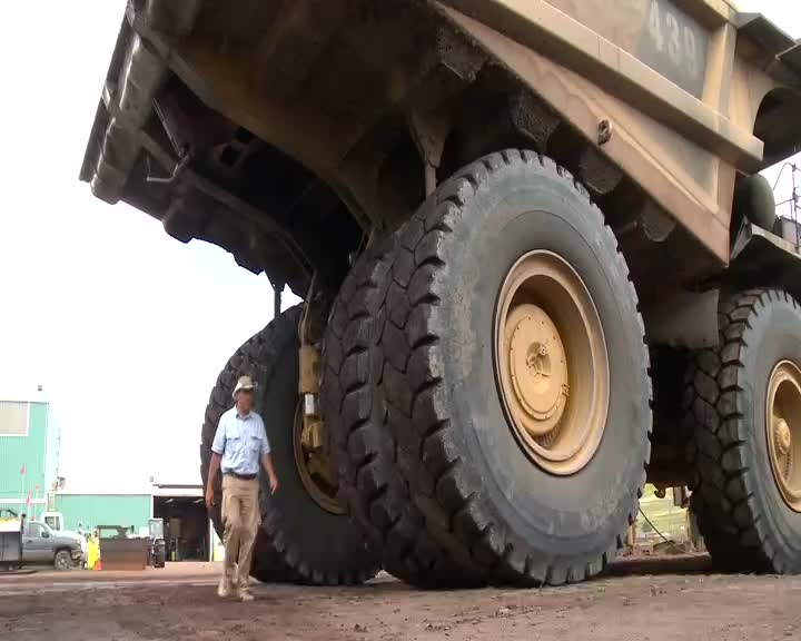 A man walks past the massive tires of a colossal yellow dump truck. The sheer scale of the vehicle dwarfs him as he strolls by.