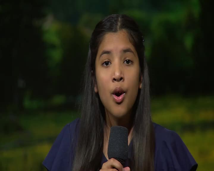 A young girl with dark hair stands before a microphone, her mouth open as if speaking. The backdrop behind her is a soft, blurred green, suggesting an outdoor or nature-themed setting.