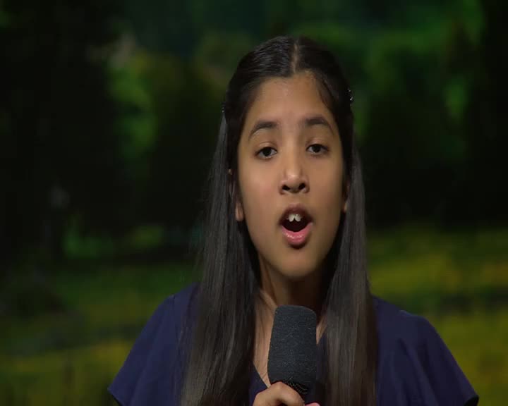 A young girl with dark hair sings into a microphone. Her mouth is open as she delivers her lines.