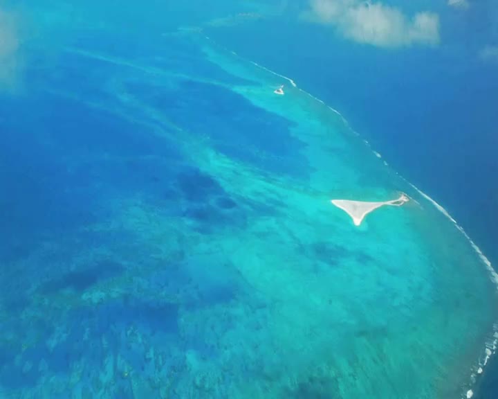 The turquoise water reveals shallow reefs and sandbars. A small, white sand island emerges from the sea.
