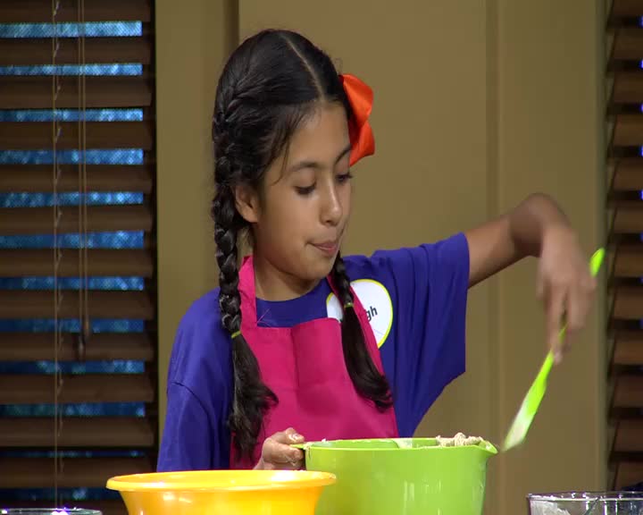 A young girl with braided hair and a bright orange bow mixes ingredients in a green bowl. She's wearing a purple shirt and a pink apron, ready for whatever 3ABN Kids has planned.
