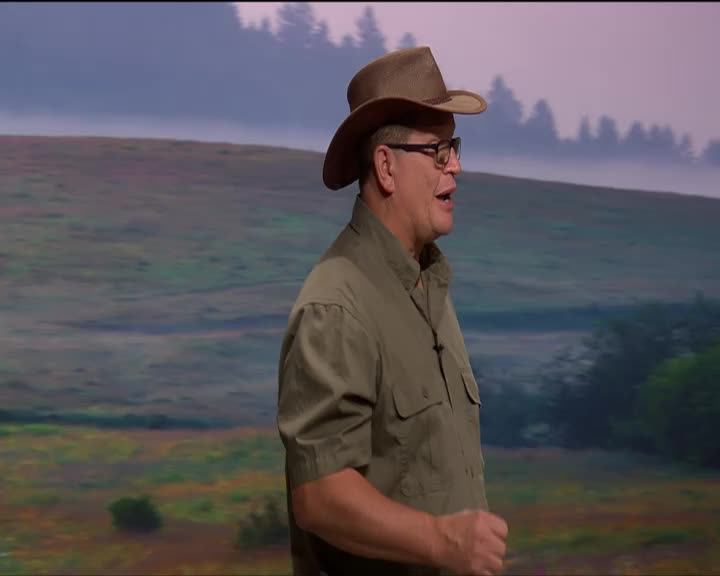 A man in a cowboy hat and a khaki shirt speaks animatedly. He stands before a painted backdrop of rolling hills and trees.