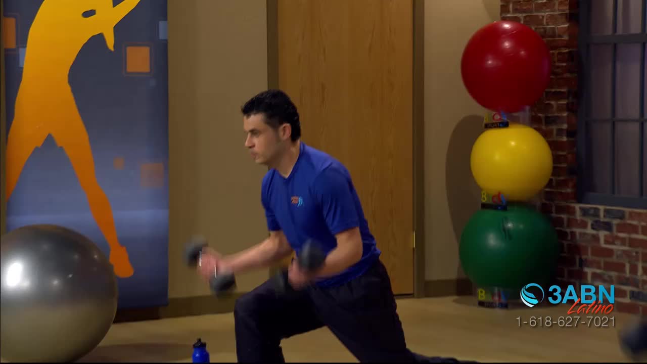 A man in a blue shirt is doing bicep curls while lunging. The set, with a 3ABN Latino logo, has exercise balls stacked in a row.
