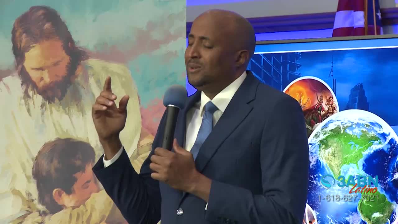 A man in a blue suit gestures with his right hand, holding a microphone as he speaks. Behind him, a large screen displays a logo for 3ABN Latino and a painting of Jesus.
