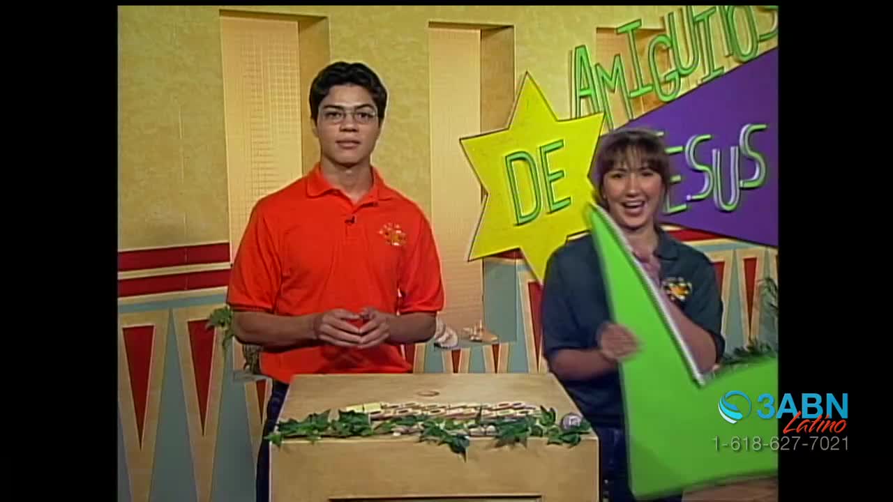 A man in an orange shirt stands beside a woman holding a large green checkmark, both smiling at the camera. Behind them, a sign in Spanish reads "Amiguitos de Jesús," and the 3ABN Latino logo is visible in the corner.

