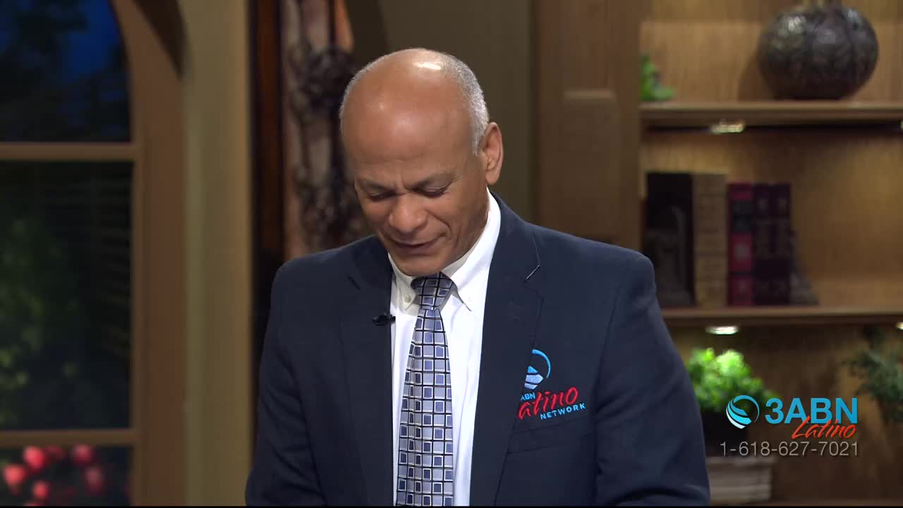 A man in a suit, with a 3ABN Latino logo on his jacket, is looking down. He appears to be reading or speaking, his expression thoughtful.
