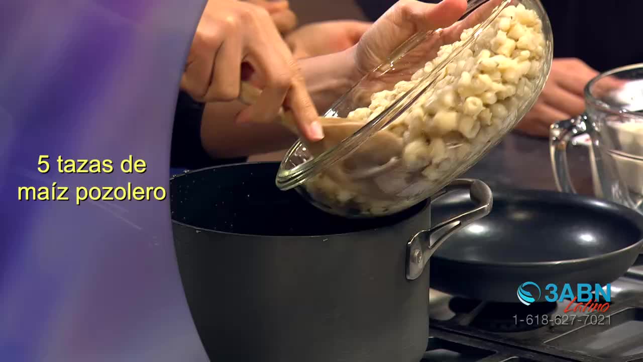 A woman is carefully pouring hominy from a glass bowl into a dark pot on the stove. The 3ABN Latino logo is visible in the corner.
