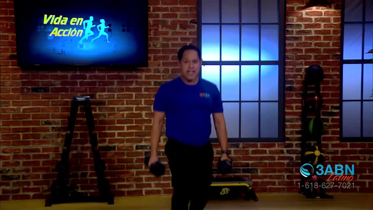 A man in a blue shirt and black pants is lifting dumbbells, his face set in concentration.  The scene unfolds in a studio, likely for a fitness program on 3ABN Latino, with a brick wall and workout equipment visible.
