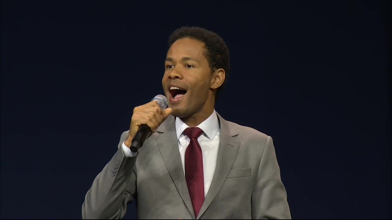 A man in a gray suit holds a microphone, his mouth open as if he's singing. He's in front of a dark blue backdrop, clearly on stage.

