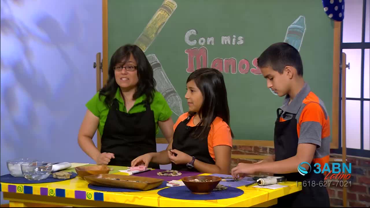 A woman in a green shirt and two children are working at a colorful table, all wearing aprons. The scene appears to be a crafting segment on 3ABN Latino, with a chalkboard in the background that says "Con mis Manos."
