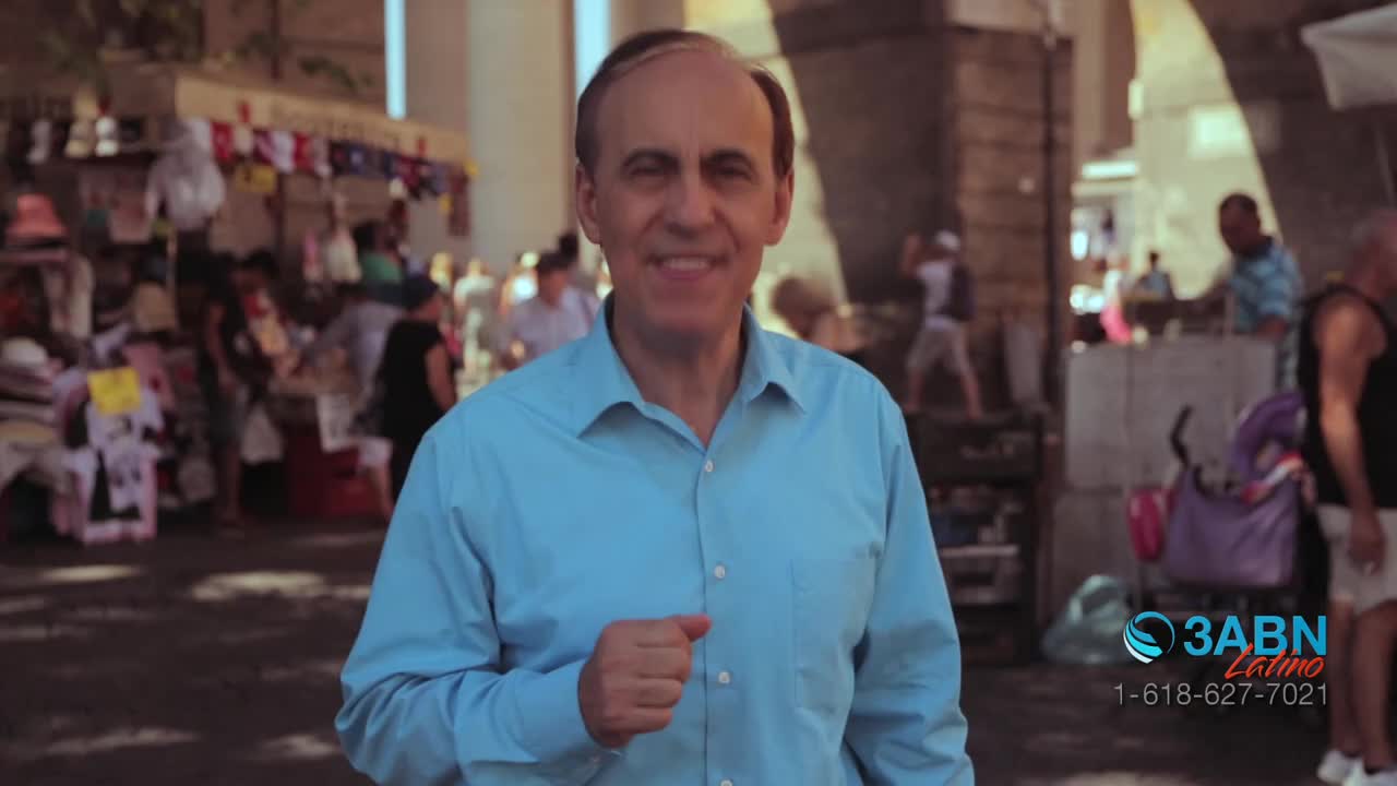 A man in a light blue shirt smiles directly at the camera, gesturing with his right hand. Behind him, a bustling outdoor market scene unfolds, with people milling about and colorful displays.
