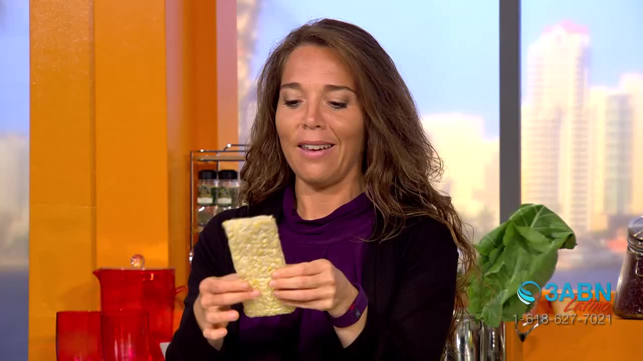 A woman in a purple top and black cardigan holds up a rectangular cracker, looking directly at it with a slight smile. The 3ABN Latino logo is visible in the corner.

