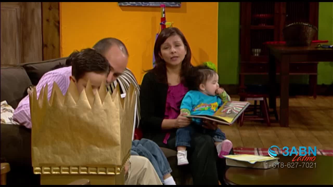 A family is gathered on a couch, with a young child in a mother's lap looking at a book. The boy in front is wearing a large golden crown.
