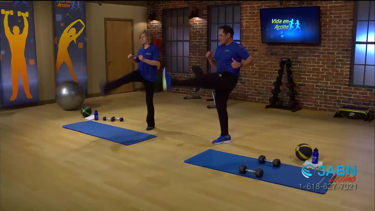 A man and a woman, both in blue shirts, are kicking their legs up high on blue mats. The scene is a fitness program from 3ABN Latino.
