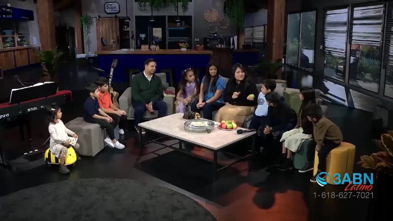 A group of people, including several children, sit on a set with a coffee table in front of them. A woman speaks into a microphone, while a small child sits on a toy bee.
