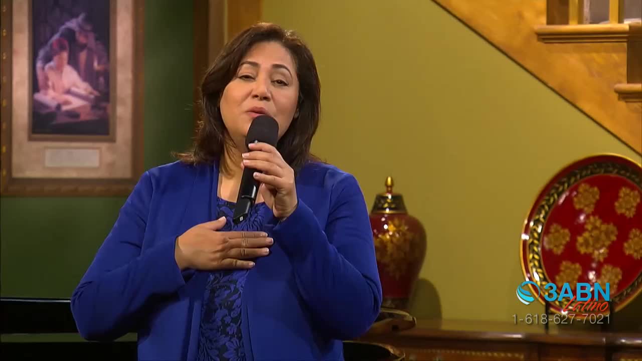 A woman in a blue jacket holds a microphone, singing with her hand over her heart. The 3ABN Latino logo is visible in the corner of the studio.

