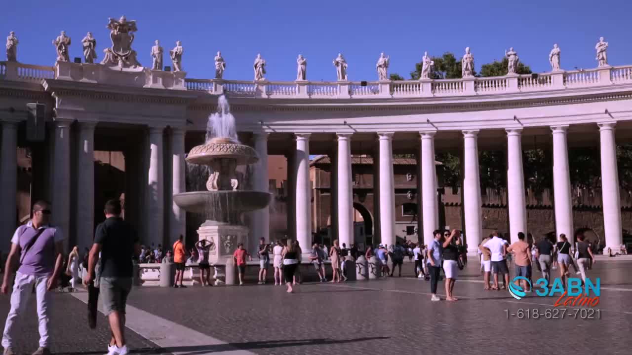 People stroll across the vast plaza, some pausing to take pictures near the fountain. The scene, broadcast by 3ABN Latino, unfolds under the colonnade's shadow.
