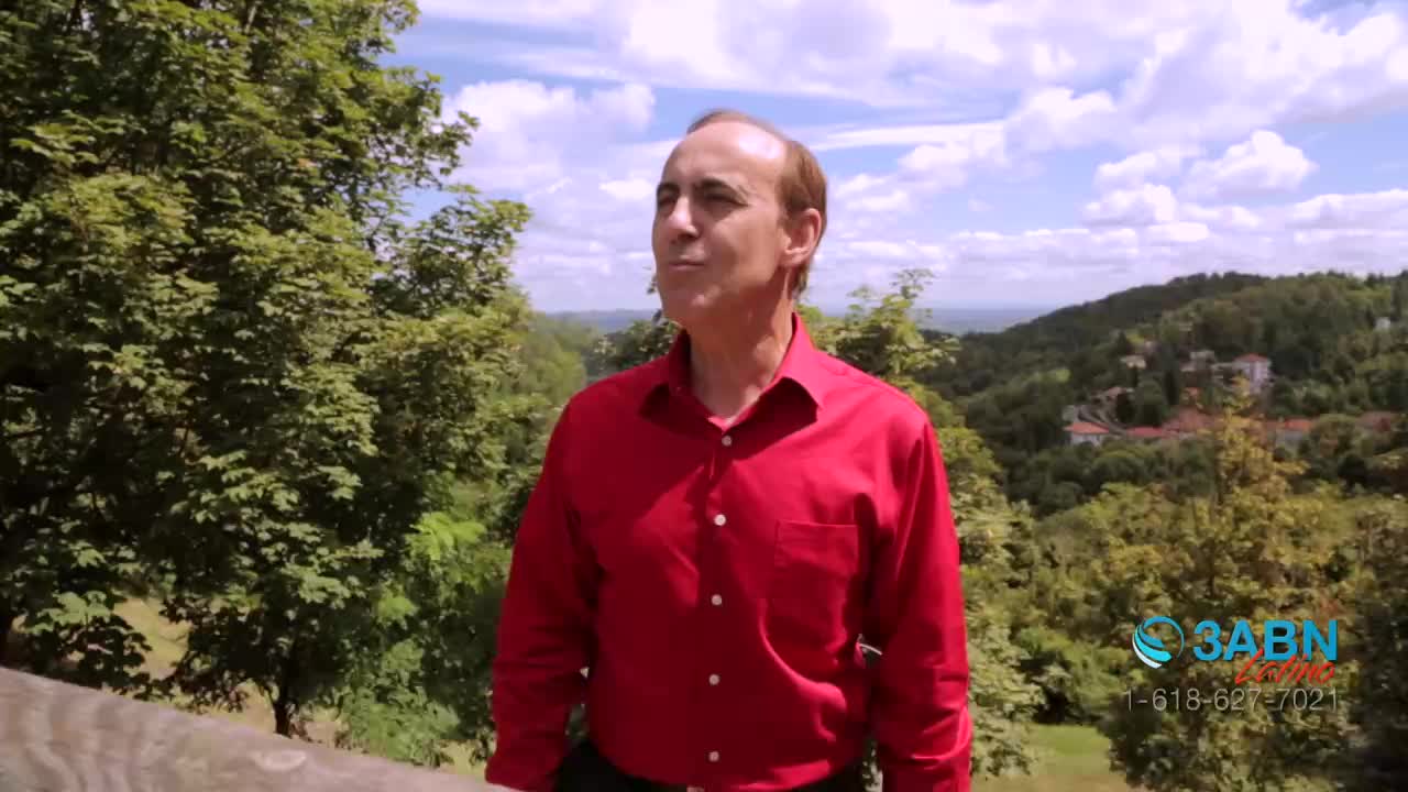 A man in a red shirt stands on a wooden ledge, looking up at the sky. Behind him, the camera captures a lush green hillside with a blue sky and fluffy white clouds.
