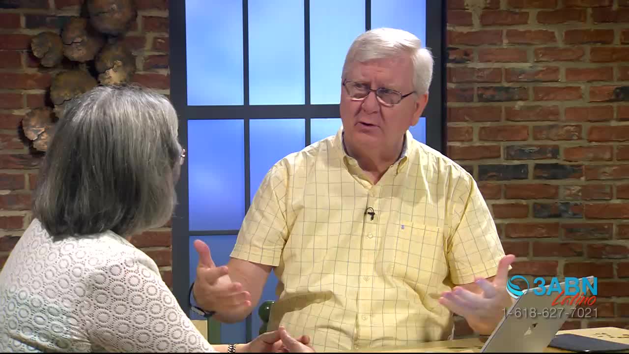 A man in a yellow shirt gestures with both hands as he speaks, his glasses reflecting the studio lights. Across from him, a woman listens intently, her grey hair framing her face.
