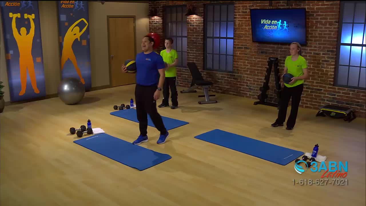 A man in blue walks toward the camera, holding a medicine ball. Two others, each with a ball, stand on mats, getting ready to exercise on a set for 3ABN Latino.
