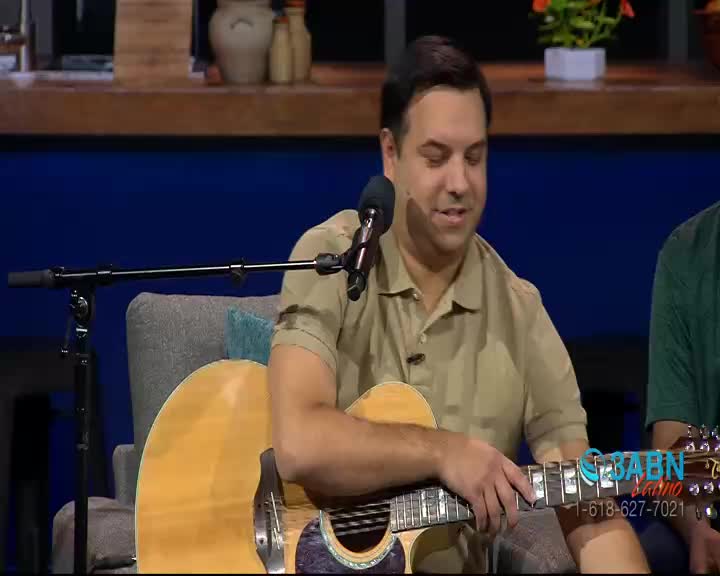 A man in a tan shirt sits with an acoustic guitar, fingers poised over the frets. He's speaking into a microphone, likely on a program for 3ABN Latino.
