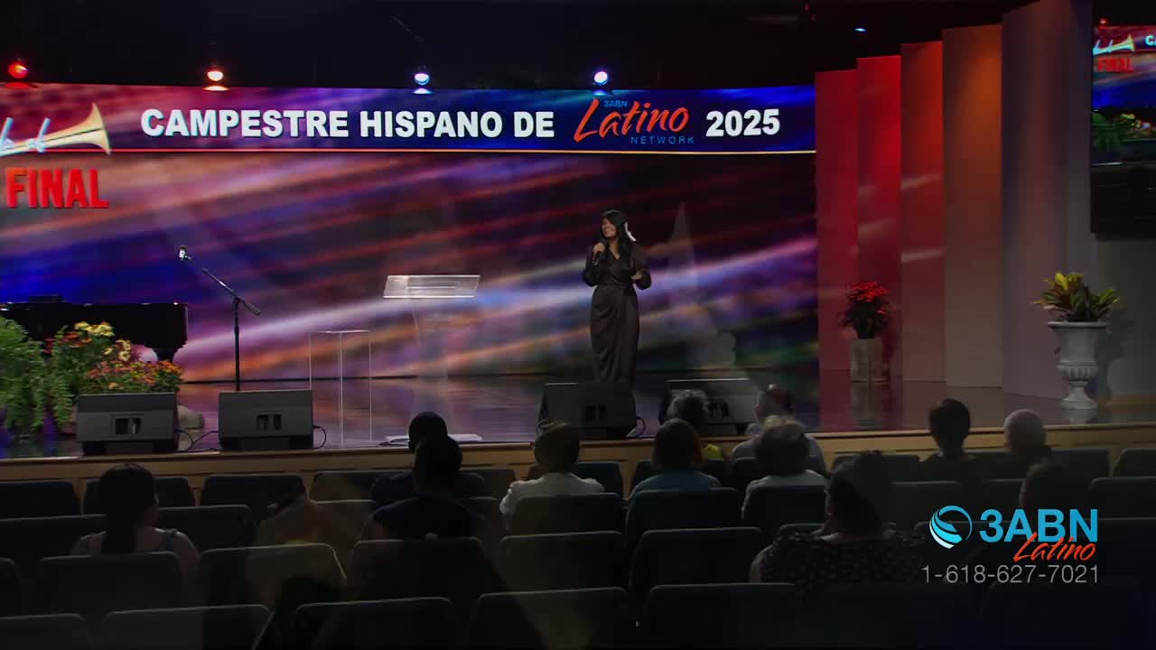A woman in a long black dress speaks into a microphone on a stage with a backdrop that says "Campestre Hispano de Latino 2025." The audience sits in rows of chairs, watching the presentation from 3ABN Latino.
