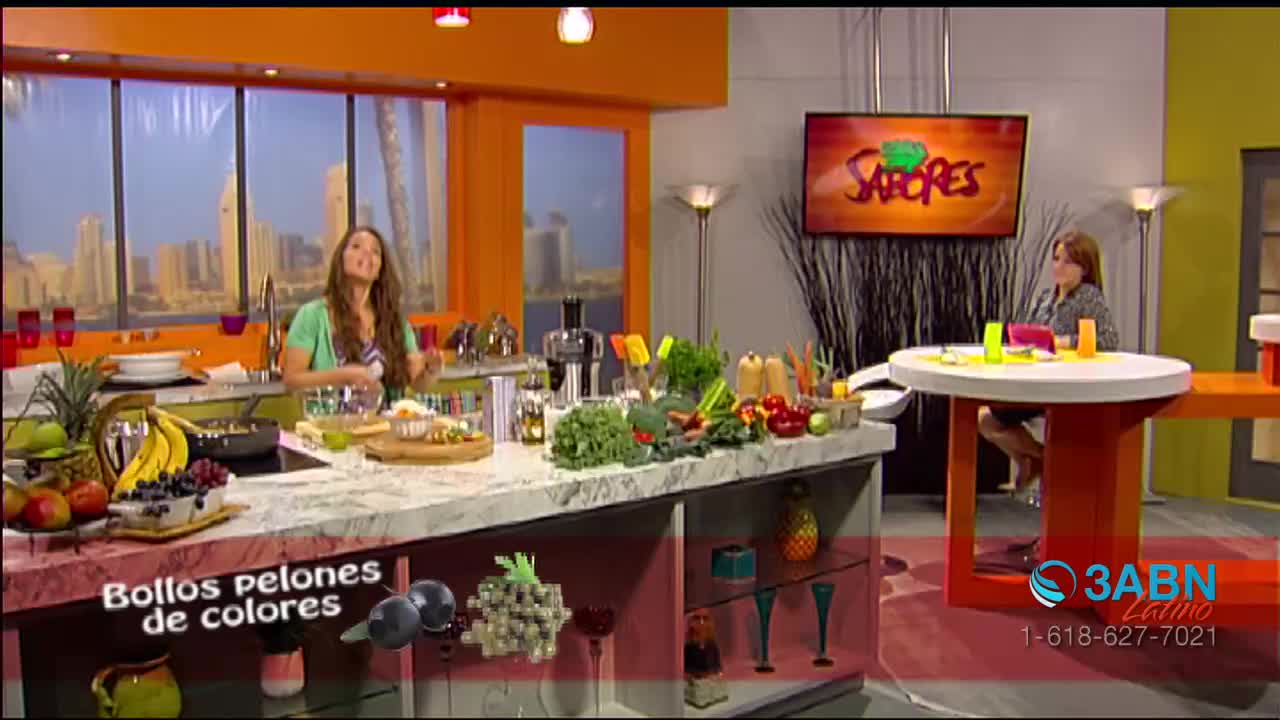 A woman in a green shirt gestures towards a countertop laden with fresh produce. Across the studio, another woman sits at a small table, while a television screen displays the show's logo.
