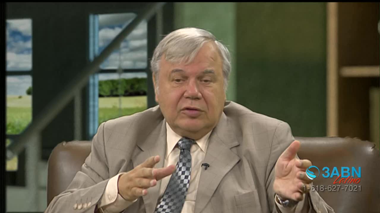 The man in the light-colored suit gestures with both hands as he speaks. Behind him, a window shows a painted backdrop of a sunny day.
