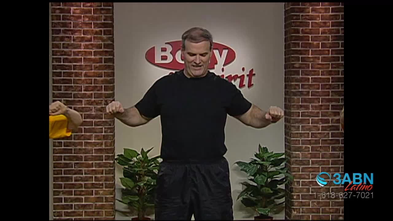 A man in a black t-shirt and shorts stands with his arms outstretched, making fists. Behind him, a brick wall and potted plants frame the "Body & Spirit" logo, all on the set of 3ABN Latino.

