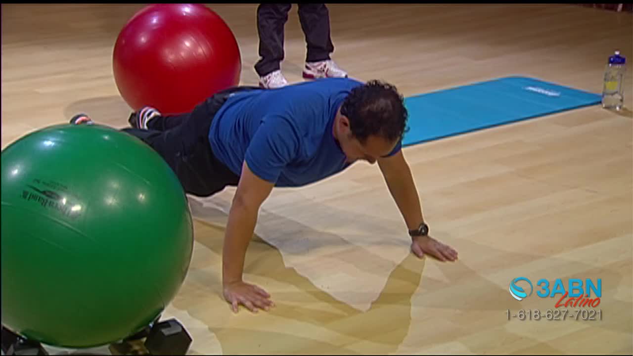 A man in a blue shirt is doing push-ups on the floor, his hands planted firmly. Behind him, someone's feet are visible, and a red and green exercise ball sit nearby.
