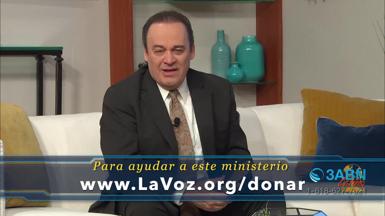 A man in a suit sits on a white couch, speaking on a 3ABN Latino program.  Text on the screen invites viewers to donate to the ministry through a website.
