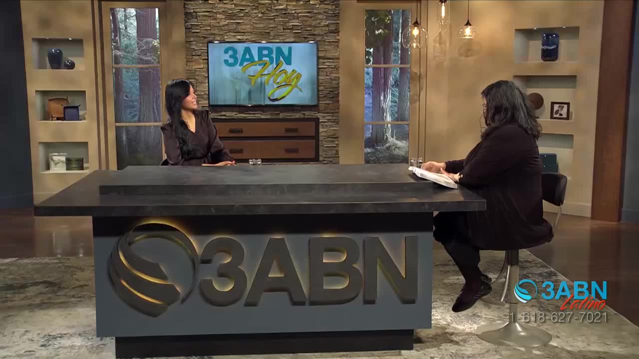 Two women sit at a large desk, one facing the camera and the other looking down at papers. Behind them, a screen displays the 3ABN Hoy logo.
