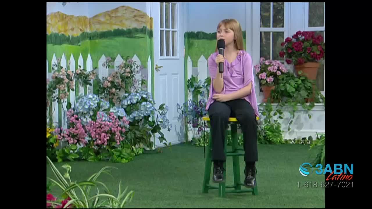 A young girl sits on a green stool, holding a microphone in front of a painted backdrop of a garden. The 3ABN Latino logo is visible in the corner.
