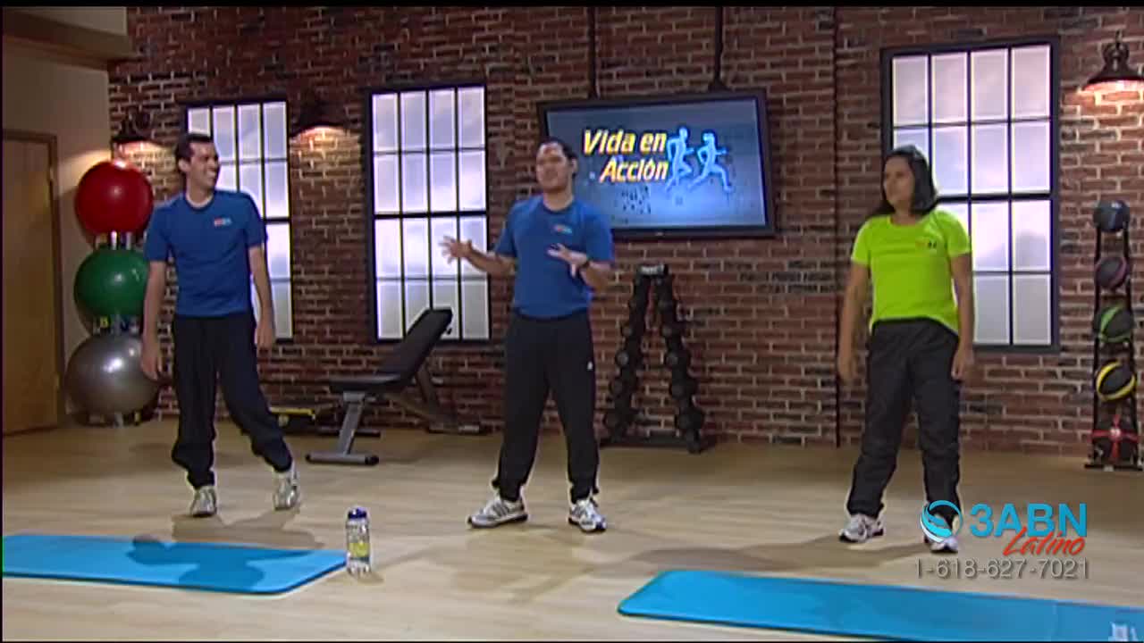 Three people are in a studio, dressed for exercise, with a "Vida en Acción" logo on a screen behind them. The man in the center gestures with his hands, while the others stand on mats, ready to move.
