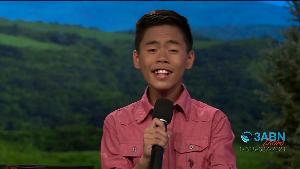 A young man in a pink shirt holds a microphone, smiling as he speaks. Behind him, a painted backdrop depicts a green landscape, and the 3ABN Latino logo is visible.

