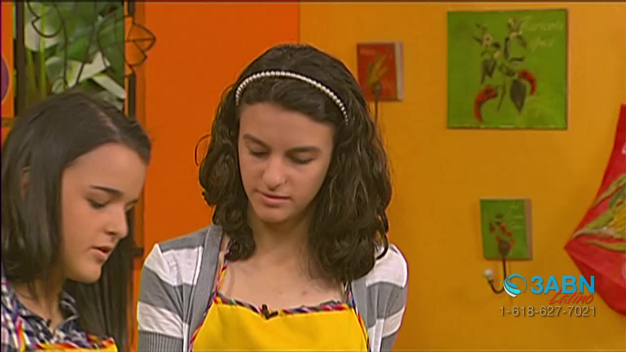 Two young women, wearing aprons, are focused on something just out of view. The scene is a brightly colored kitchen set, possibly for a program on 3ABN Latino.
