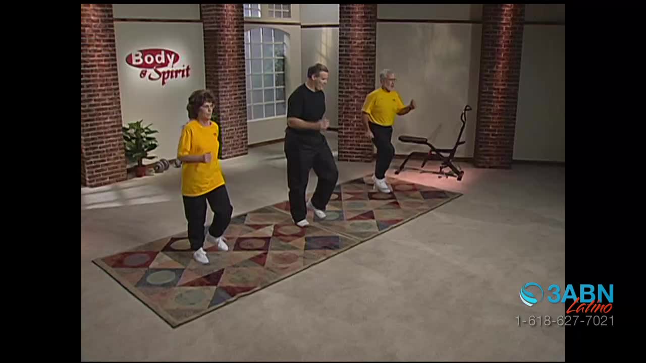 Three people in workout clothes are jogging in place on a patterned rug. The woman in the yellow shirt and black pants is leading the exercise.
