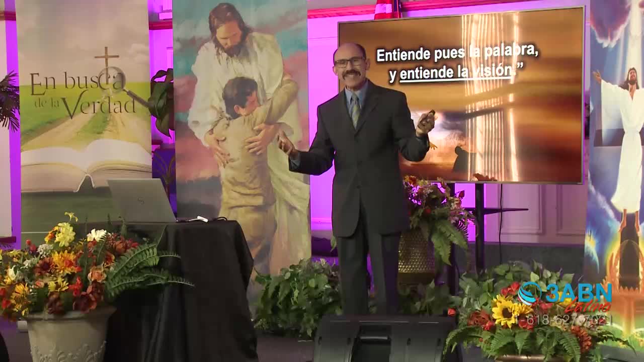 A man in a dark suit gestures with both hands, speaking in front of a screen displaying Spanish text. Behind him, colorful banners and floral arrangements decorate the stage for a 3ABN Latino broadcast.
