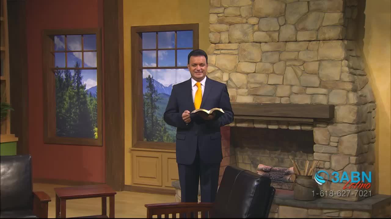 A man in a suit stands in front of a stone fireplace, holding an open book. He faces the camera, smiling, with a backdrop of painted windows and a 3ABN Latino logo in the corner.
