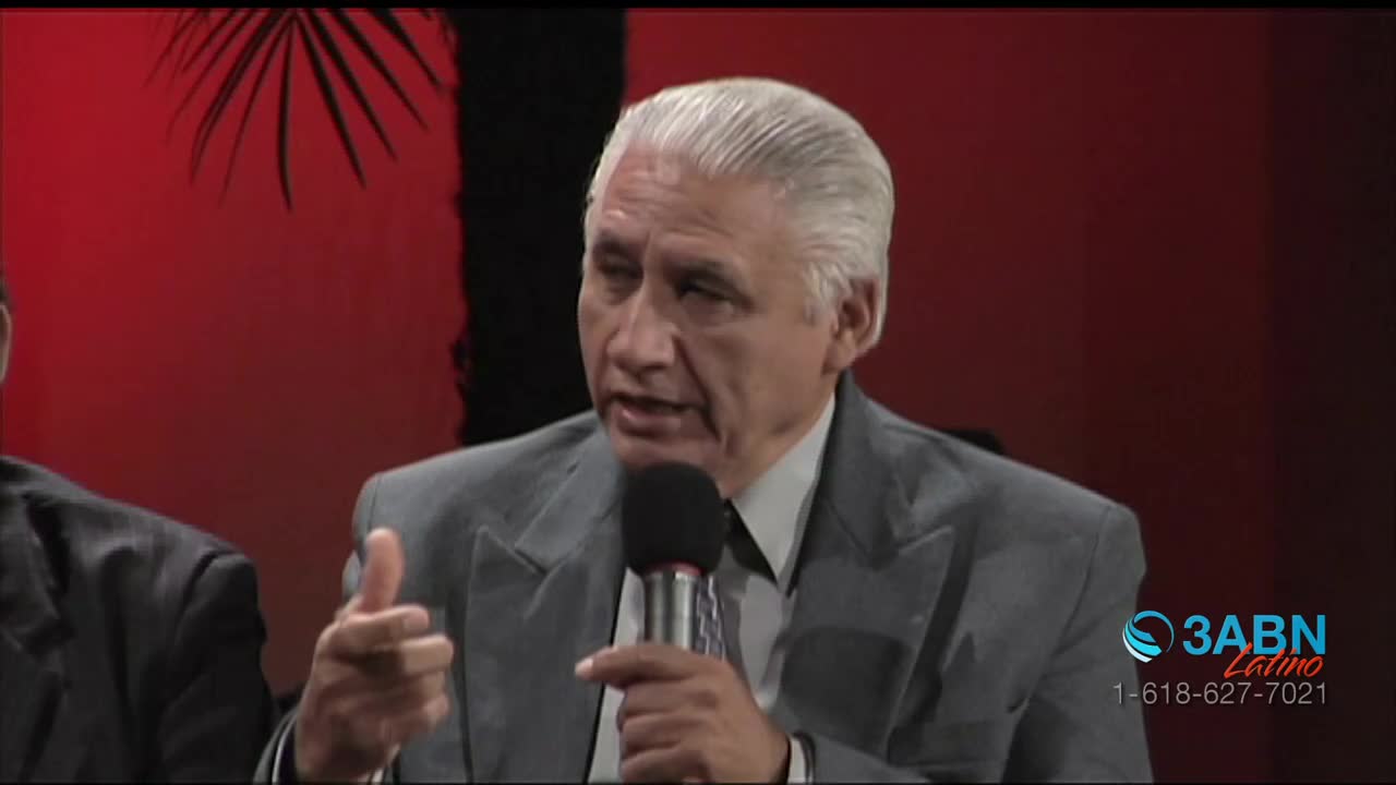 A man in a gray suit speaks into a microphone, gesturing with his left hand. Behind him, a red backdrop and the 3ABN Latino logo are visible.
