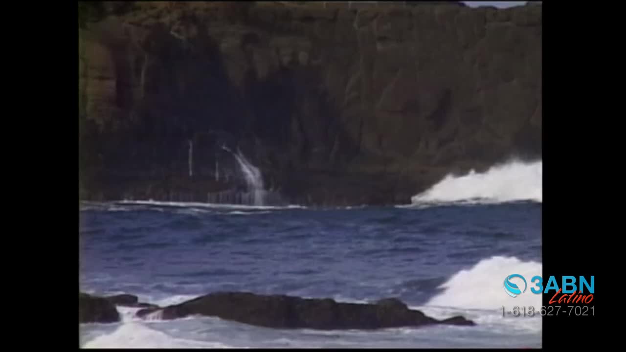 Water crashes against dark rocks, creating white foam. A small waterfall trickles down the cliff face.
