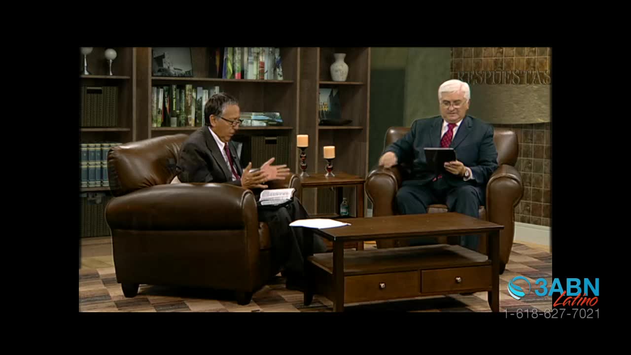 Two men in suits sit in leather armchairs, facing each other in a studio. One gestures with his hands while the other holds a tablet, both appearing to be engaged in a discussion for 3ABN Latino.
